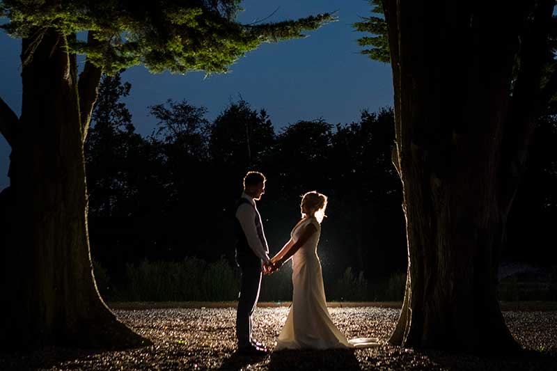 Bride and Groom in Silhouette at Night Standing Between Two Large Oak Trees 